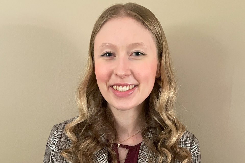 Abigail Smith, with long blond hair, wearing a flannel professional jacket, smiles and stands in front of a beige wall.